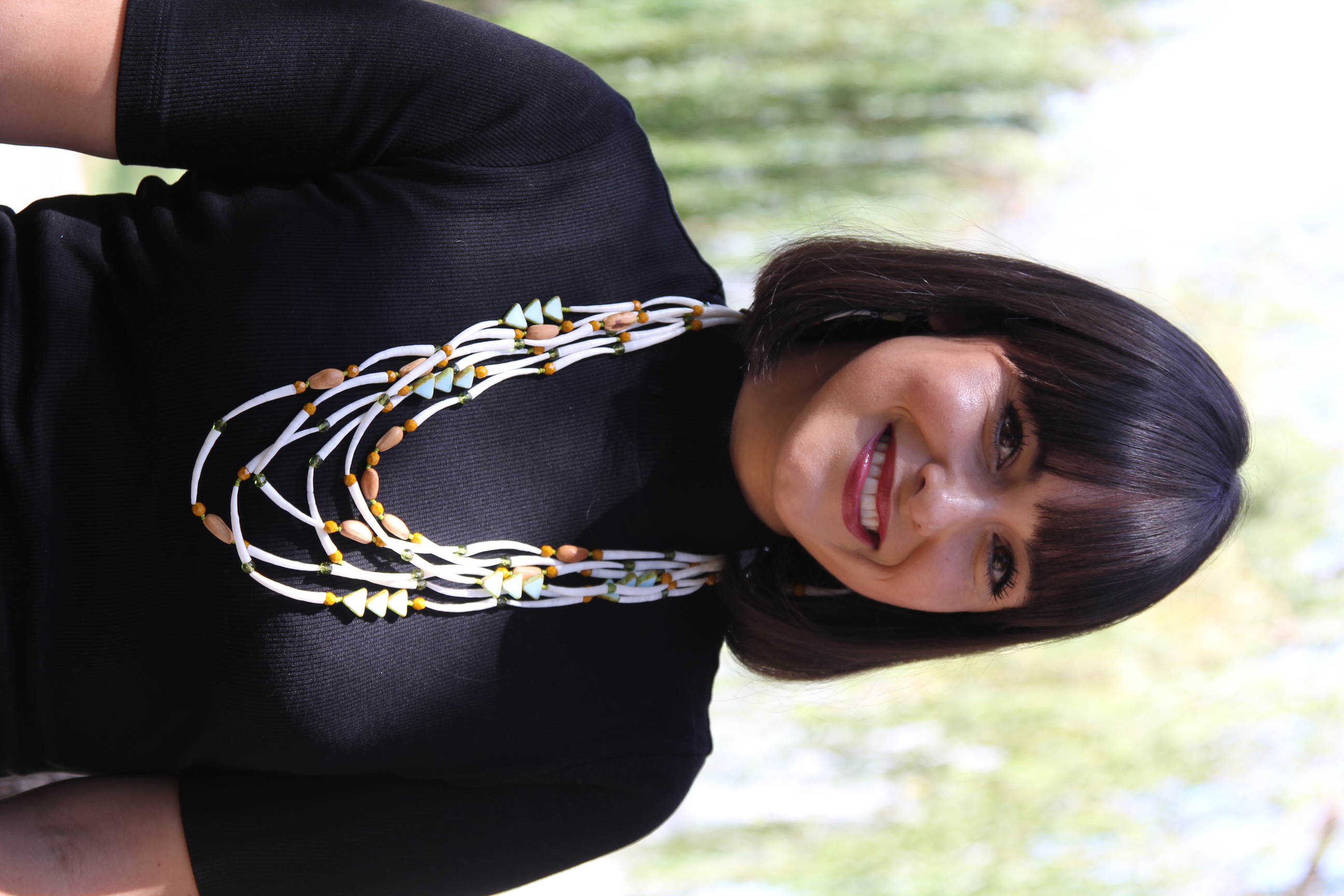 Photo portrait of a woman standing outdoors; she is smiling and wears a black top and a beaded necklace with many strands