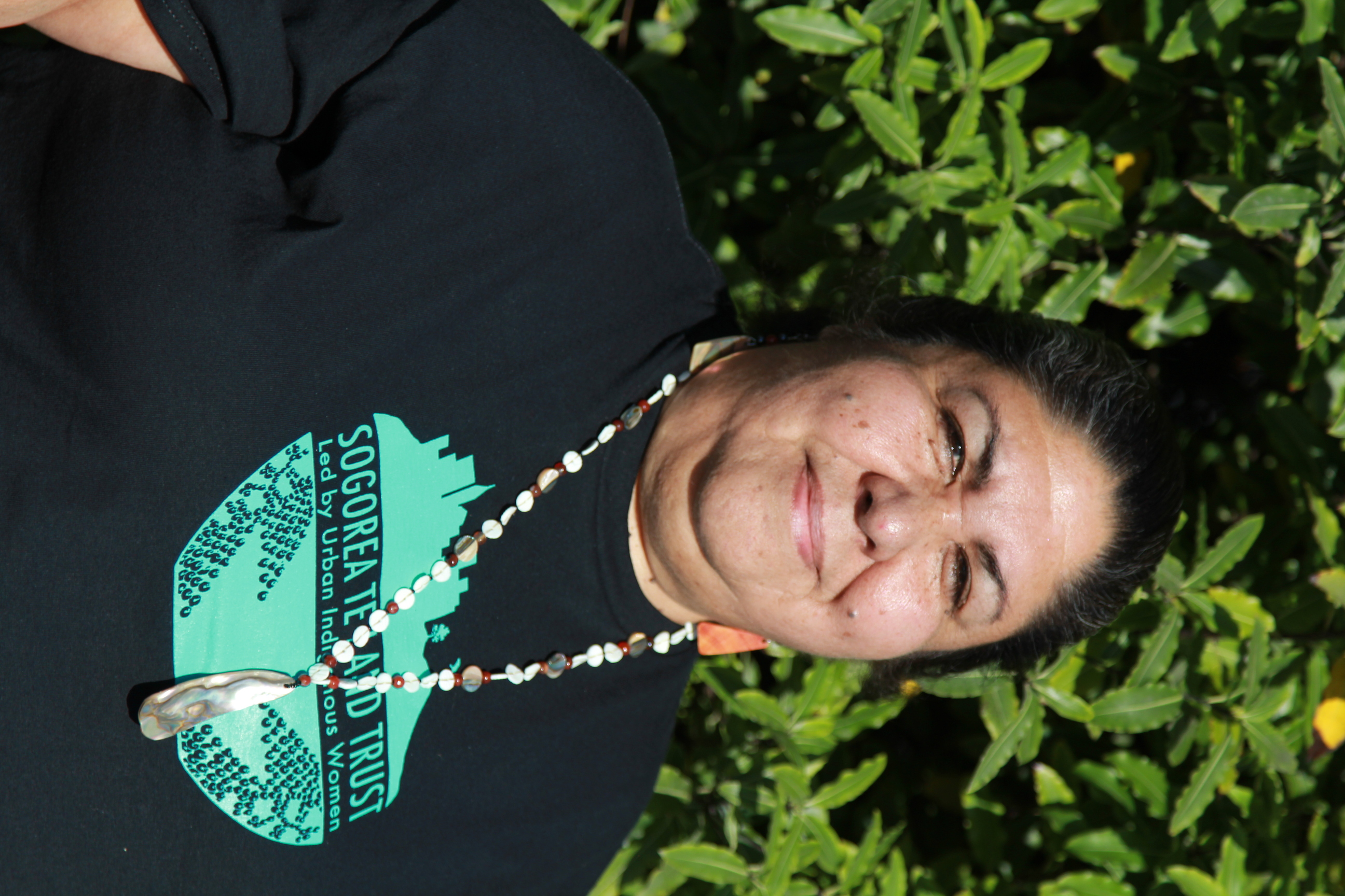 Photo portrait of a woman standing outdoors; she wears a dark t-shirt that read Sogorea Te Land Trust and a beaded necklace