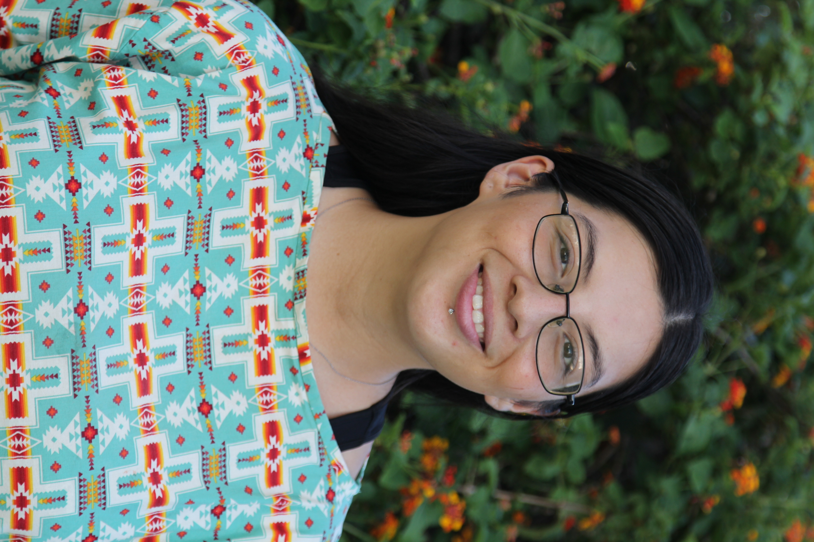 Photo portrait of a woman standing outdoors, she is smiling and wears a turquoise top patterned with white, red, gold and purple