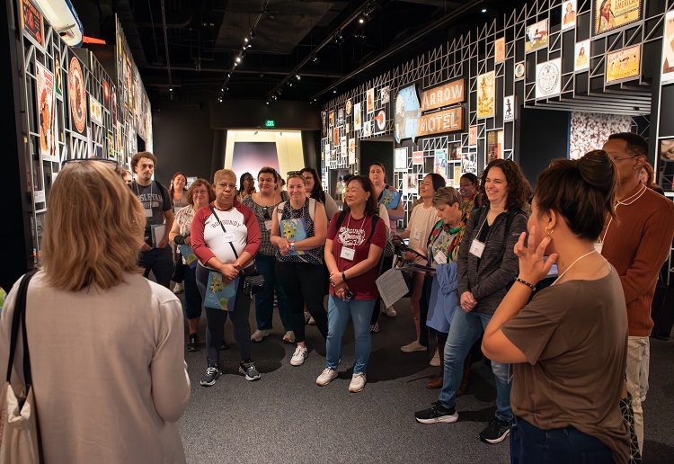 a large, diverse group of people gathered in a museum exhibition with exhibit graphics in the background