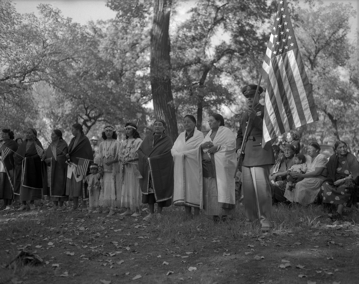 Pascal Cleatus Poolaw Sr., holding the American flag, with members of the Kiowa War Mothers outside in Oklahoma