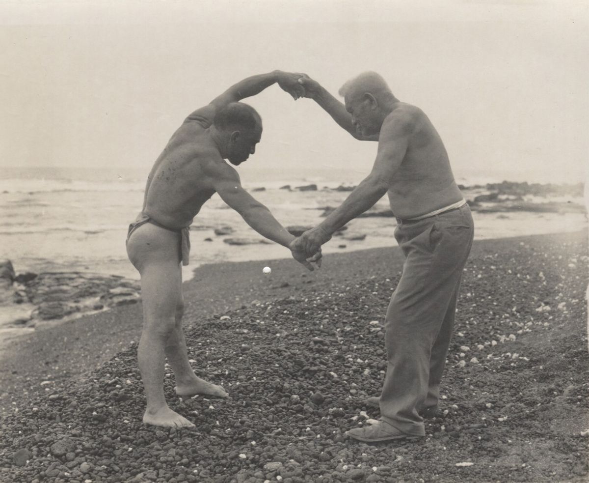 Two native Hawaiian men demonstrate traditional martial arts by holding hands and streching arms in a circle on the beach